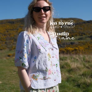 May include: A light blue button-down shirt with a wide collar and short puff sleeves, featuring colorful floral embroidery. The shirt displays the text 'jen byrne' and 'Studio Nine' in white script. It is paired with floral print pants.