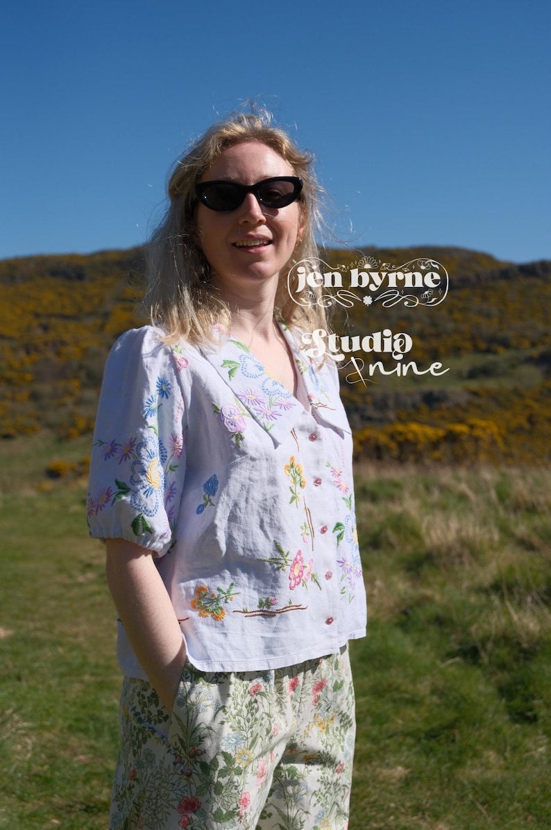 May include: A light blue button-down shirt with a wide collar and short puff sleeves, featuring colorful floral embroidery. The shirt displays the text 'jen byrne' and 'Studio Nine' in white script. It is paired with floral print pants.