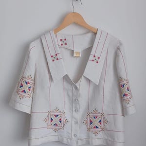 May include: A white short-sleeved button-down shirt featuring a large collar and geometric embroidery in red, blue, and gold thread. The shirt includes the text "made by Lorraine using the Tilly Pattern" at the bottom of the image.