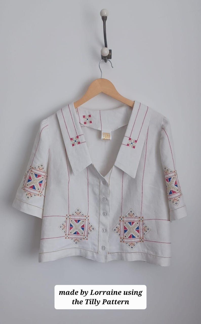 May include: A white short-sleeved button-down shirt featuring a large collar and geometric embroidery in red, blue, and gold thread. The shirt includes the text "made by Lorraine using the Tilly Pattern" at the bottom of the image.