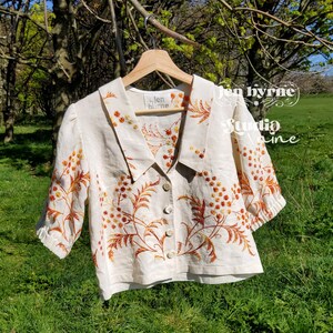 May include: A cream-colored short-sleeve blouse featuring a wide collar, a front button closure, and intricate orange and yellow floral embroidery. The garment includes a label reading "jen byrne" and is displayed on a wooden hanger.