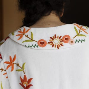 May include: A white jacket featuring a wide collar and fabric surface embroidered with floral patterns in shades of orange, red, yellow, and green. The embroidery includes stylized flowers and leaves, with a decorative zigzag stitch line.