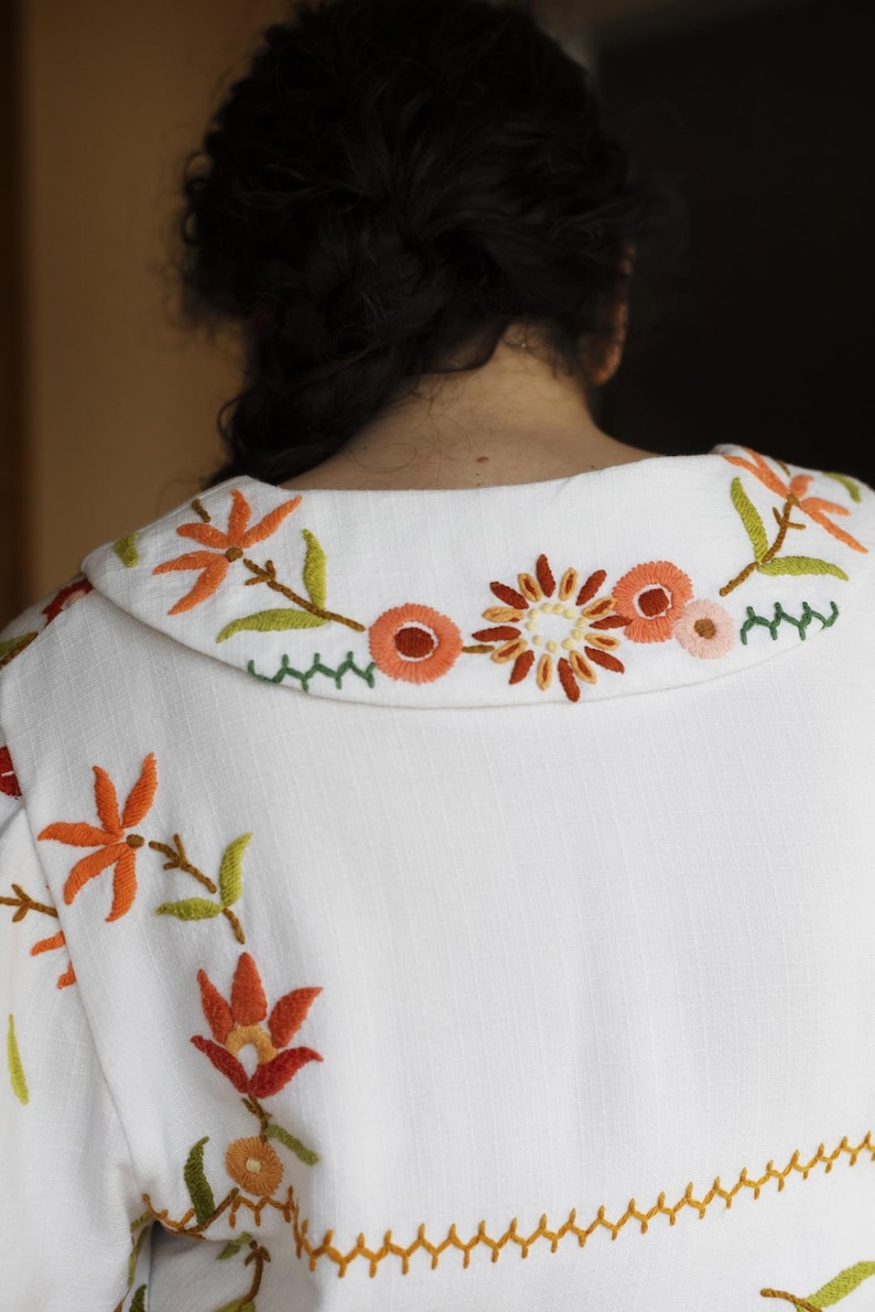 May include: A white jacket featuring a wide collar and fabric surface embroidered with floral patterns in shades of orange, red, yellow, and green. The embroidery includes stylized flowers and leaves, with a decorative zigzag stitch line.