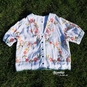 May include: A white short-sleeved blouse featuring a V-neckline with a wide collar, a button-down front, and colorful floral embroidery in shades of pink, orange, yellow, and green. The label inside reads Jen Byrne, and the bottom hem is green.