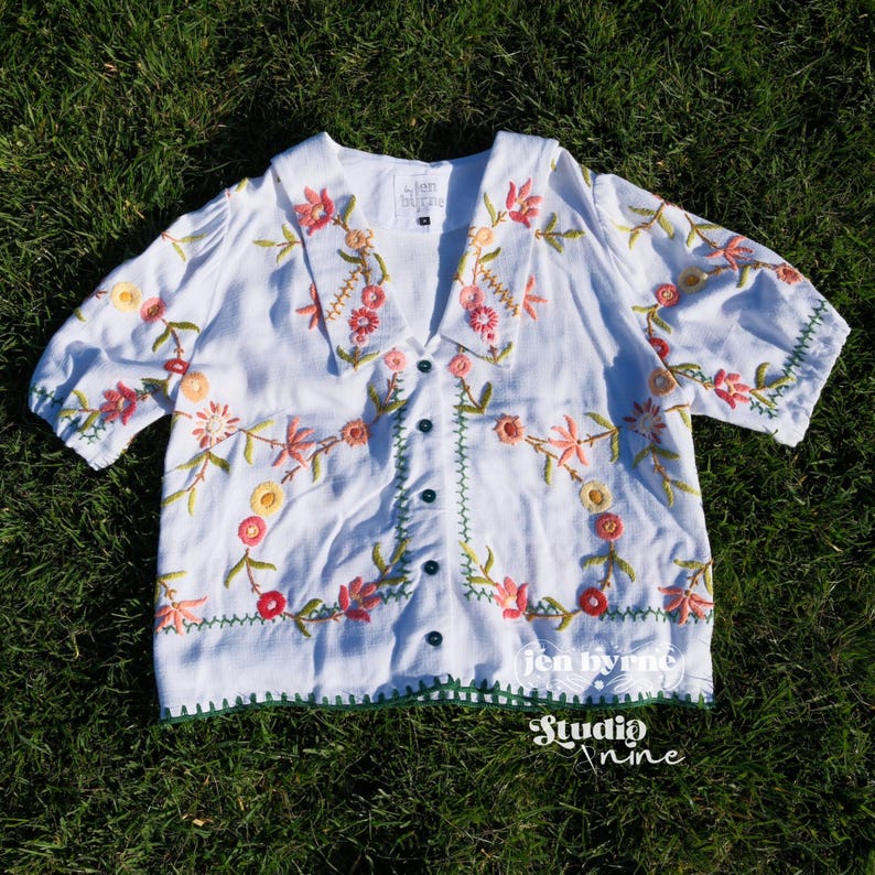May include: A white short-sleeved blouse featuring a V-neckline with a wide collar, a button-down front, and colorful floral embroidery in shades of pink, orange, yellow, and green. The label inside reads Jen Byrne, and the bottom hem is green.