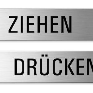 Op de afbeelding: Twee zilveren metalen borden met zwarte tekst. Het bovenste bord zegt "ZIEHEN" en het onderste bord zegt "DRÜCKEN".