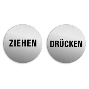 Puede incluir: Dos letreros redondos de metal plateado con texto negro. El letrero de la izquierda dice "ZIEHEN" y el letrero de la derecha dice "DRÜCKEN".