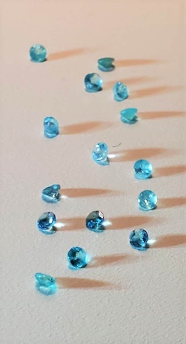 May include: A collection of 15 light blue gemstones scattered on a white surface.