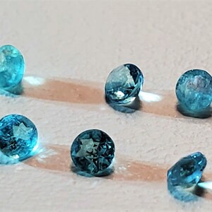 May include: Six round, faceted, blue gemstones on a white surface. The gemstones are translucent and have a slight sparkle.