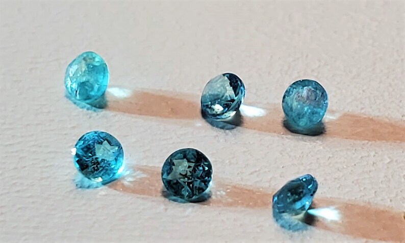 May include: Six round, faceted, blue gemstones on a white surface. The gemstones are translucent and have a slight sparkle.