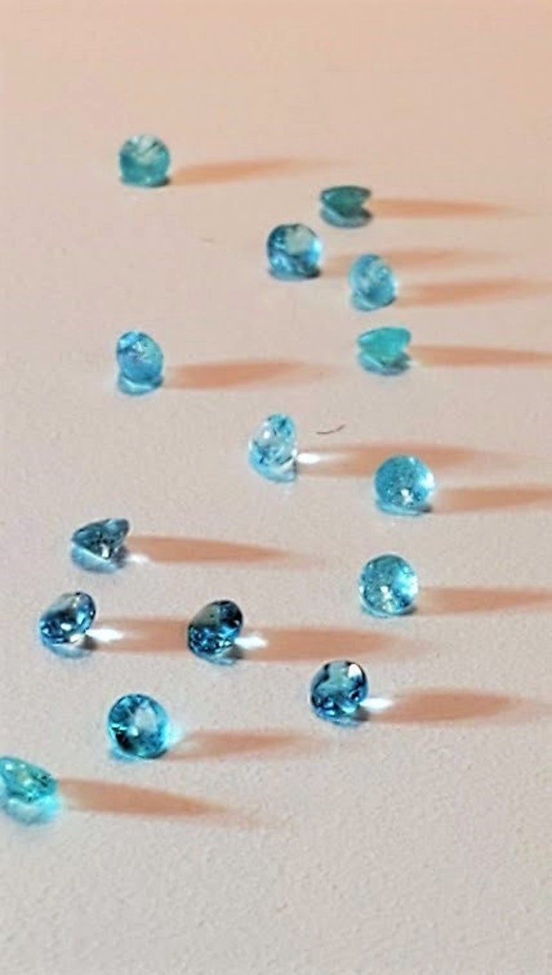 May include: A collection of small, round, light blue gemstones arranged on a white surface. The gemstones are scattered in a random pattern.