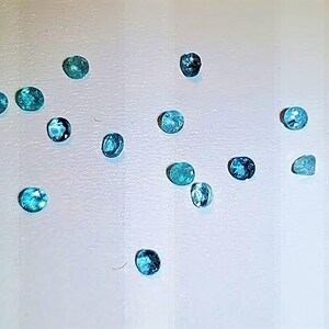 May include: A collection of 14 oval-shaped gemstones in various shades of blue and green. The gemstones are arranged on a white surface.