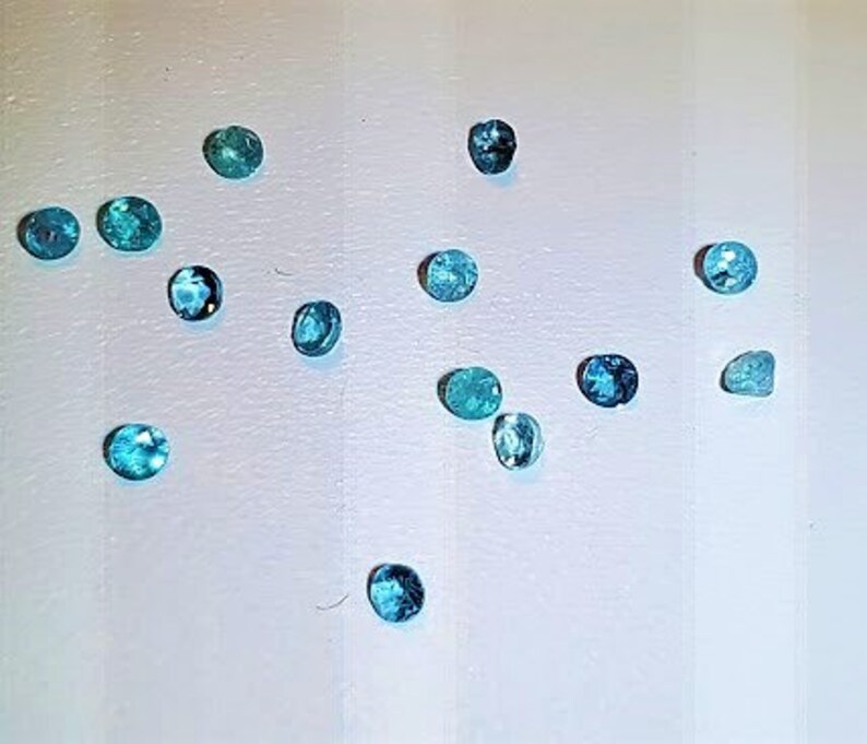 May include: A collection of 14 oval-shaped gemstones in various shades of blue and green. The gemstones are arranged on a white surface.