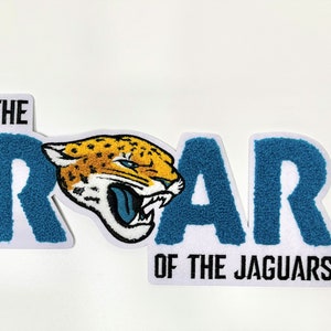 May include: A patch with the text "THE ROAR OF THE JAGUARS". The word "ROAR" is in teal, textured letters. A jaguar head graphic is in the center, with a yellow and black spotted pattern and a blue tongue.
