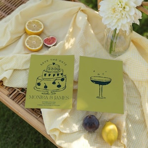 Può includere: Due biglietti verde oliva di salvataggio della data con illustrazioni blu scuro di una torta e un bicchiere da cocktail. La carta a sinistra ha il testo "SAVE THE DATE" e "MONICA & JAMES". L'immagine include anche limoni, un fico e un vaso di fiori bianchi.