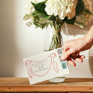 May include: A white envelope featuring a red line drawing of two birds holding a banner with handwritten address text, accompanied by three postage stamps. The envelope is positioned in front of a clear glass vase filled with white hydrangea flowers.
