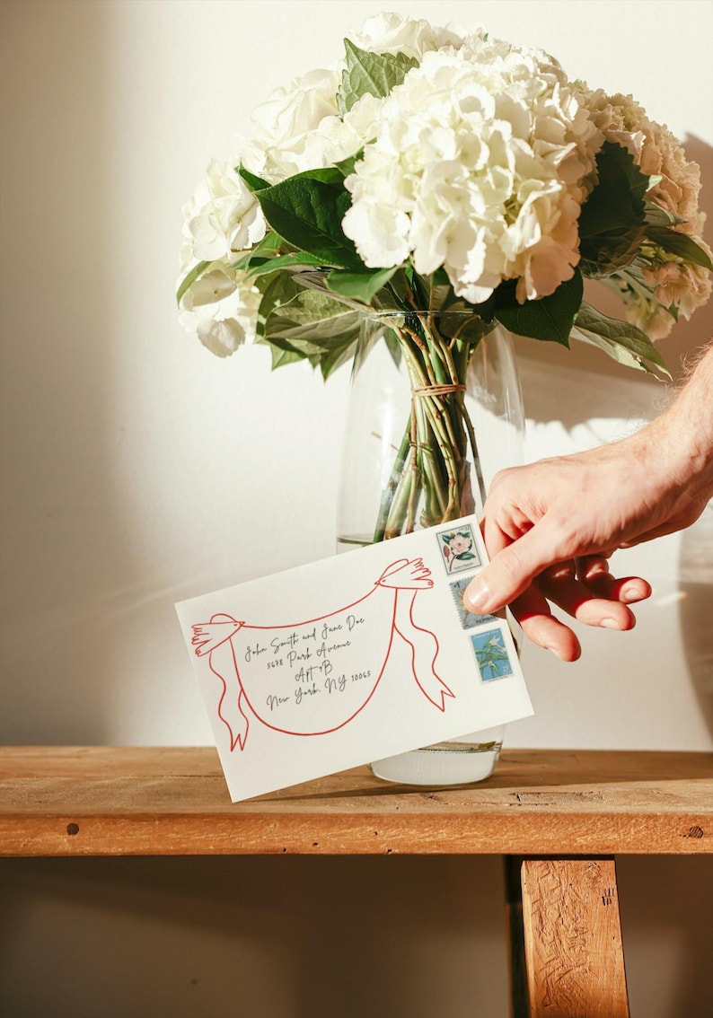 May include: A white envelope featuring a red line drawing of two birds holding a banner with handwritten address text, accompanied by three postage stamps. The envelope is positioned in front of a clear glass vase filled with white hydrangea flowers.