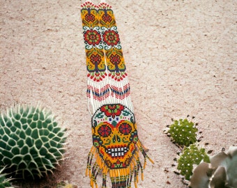 May include: A long beaded necklace with a colorful skull design and fringe. The necklace is made of white, yellow, red, blue, and green beads. The skull is surrounded by flowers and has a yellow background. The necklace is hanging against a sandy background.