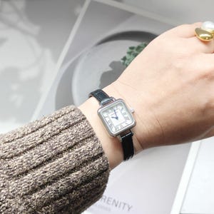 May include: A silver-toned square watch with a white face and black Roman numerals. The watch has a black leather band. The watch is on a wrist with a brown sweater.