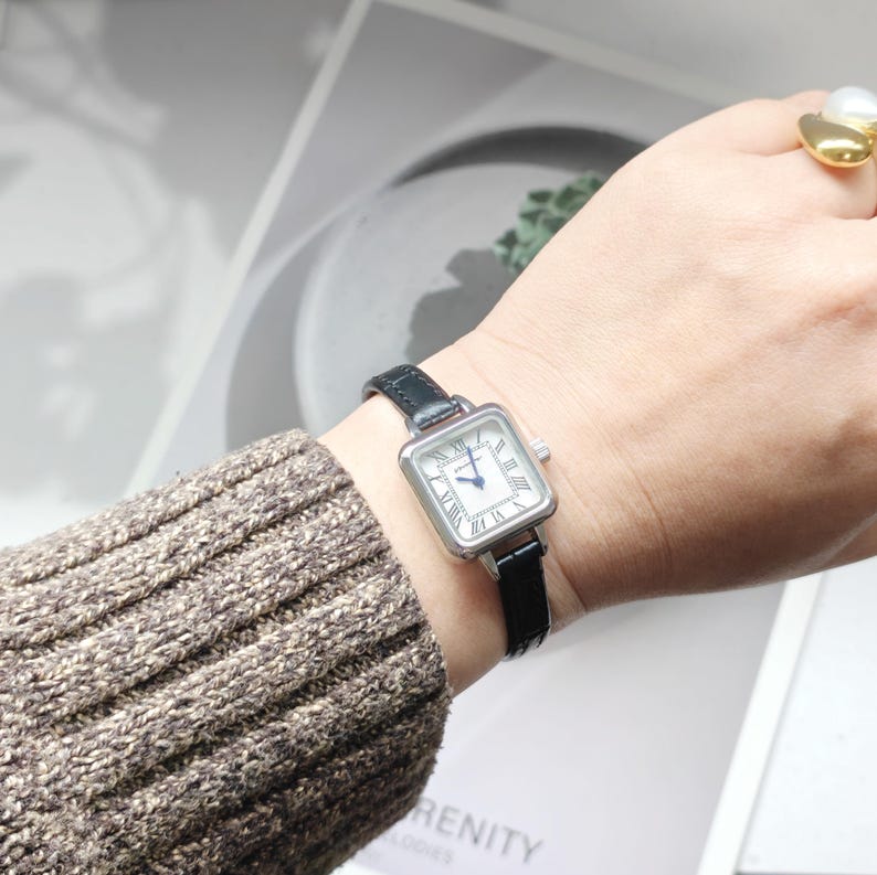 May include: A silver-toned square watch with a white face and black Roman numerals. The watch has a black leather band. The watch is on a wrist with a brown sweater.