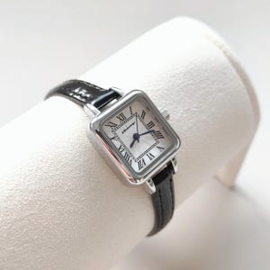 May include: A square-faced wristwatch with a silver-tone case and a black leather band. The watch face is white with black Roman numerals and blue hands. The watch is displayed on a white surface.