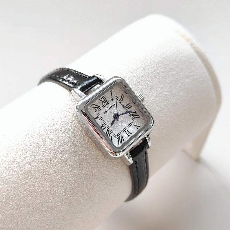 May include: A square-faced wristwatch with a silver-tone case and a black leather band. The watch face is white with black Roman numerals and blue hands. The watch is displayed on a white surface.
