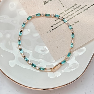 May include: A gold-toned bracelet with alternating rectangular teal and clear, round-cut gemstones. The bracelet is displayed on a white plate with a gold rim, next to a card with text. The text on the card reads "greatest happiness in life".