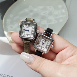 May include: Two square-faced wristwatches with Roman numeral hour markers and a small seconds hand. One watch has a light brown leather band, and the other has a black leather band. The watch faces are white with a silver-colored case.