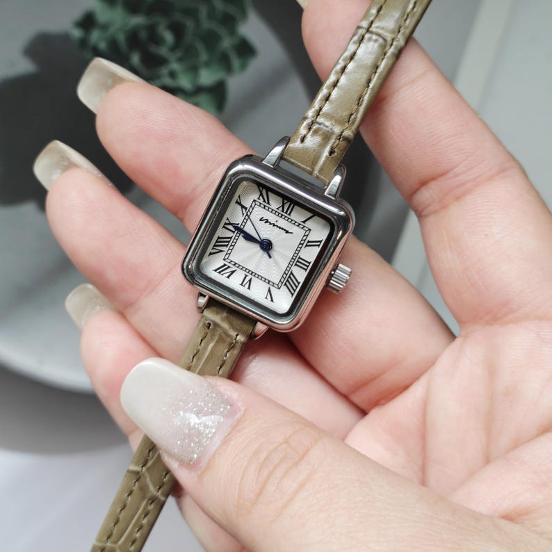 May include: A square-faced wristwatch with a silver-colored case and a light-colored leather band. The watch face is white with black Roman numerals and black hands. The band is approximately 0.5 inches wide.