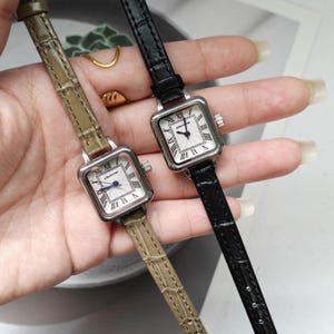 May include: Two square-faced wristwatches with silver-toned cases and Roman numeral hour markers. One watch has a black leather band, the other a khaki-colored band. The watch faces are white with black hands. The watches are displayed side by side.