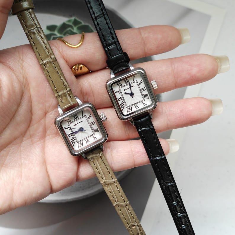 May include: Two square-faced wristwatches with silver-toned cases and Roman numeral hour markers. One watch has a black leather band, the other a khaki-colored band. The watch faces are white with black hands. The watches are displayed side by side.