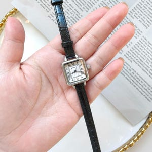 May include: A square-faced wristwatch with a black leather band. The watch has a silver-colored case and a white face with black Roman numerals. The watch is a classic design, suitable for everyday wear.