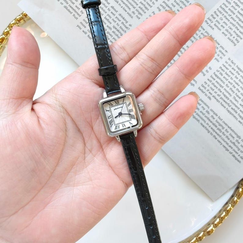 May include: A square-faced wristwatch with a black leather band. The watch has a silver-colored case and a white face with black Roman numerals. The watch is a classic design, suitable for everyday wear.