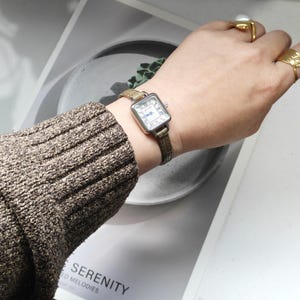 May include: A square-faced wristwatch with a silver case and a white dial featuring Roman numerals and blue hands. The watch has a brown leather band. The watch is on a wrist with a brown sweater.