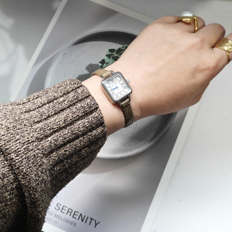 May include: A square-faced wristwatch with a silver case and a white dial featuring Roman numerals and blue hands. The watch has a brown leather band. The watch is on a wrist with a brown sweater.
