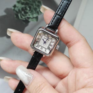 May include: A square-faced wristwatch with a silver-tone case and a black leather band. The watch face is white with black Roman numerals and delicate blue hands. The watch is held in a hand.