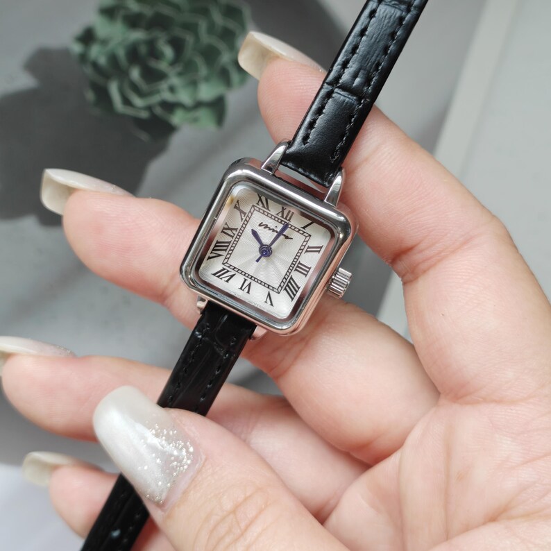 May include: A square-faced wristwatch with a silver-tone case and a black leather band. The watch face is white with black Roman numerals and delicate blue hands. The watch is held in a hand.