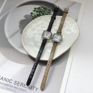 May include: Two square-faced wristwatches with leather bands. One watch has a black band, the other a tan band. Both watches have silver-colored cases and white faces with blue hands and Roman numeral hour markers. The watches are displayed on a white plate.