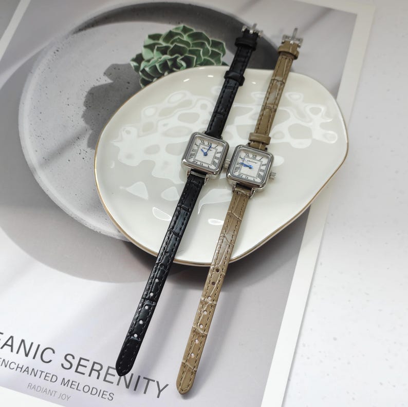 May include: Two square-faced wristwatches with leather bands. One watch has a black band, the other a tan band. Both watches have silver-colored cases and white faces with blue hands and Roman numeral hour markers. The watches are displayed on a white plate.