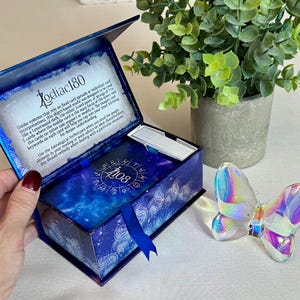 May include: A blue box with a gold ribbon and a deck of cards inside. The box has a star pattern and the text "Zodiac180" on the front. The text inside the box reads "Unlike systems that rely on fixed card spreads or individual card interpretations, this object-based oracle was designed to be read like a Lenormand deck. Use either 2 card pairs, or strings of 3 to 5 cards where the middle card is the main focus of the query and the cards on either side support or modify it. The imagery and keywords on each card will help you tap into your inner knowing for deeper understanding. Use the Astrological Archetypes when you need the people element in your reading or want advice on what characteristics you should embody when dealing with a particular situation."