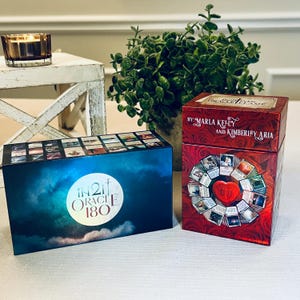 May include: Two boxes of oracle cards. The box on the left is blue with a white moon and the text "In 2 It Oracle 180". The box on the right is red with a heart and the text "By Marla Kelly and Kimberley Aria".