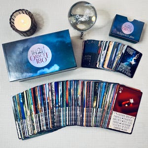 May include: A deck of 180 oracle cards with a blue box and a guidebook. The cards feature colorful images and text. The box has the text "IN21 ORACLE 180" on it. The guidebook has the text "IN21 ORACLE by KEDERLEY ARIA" on it.