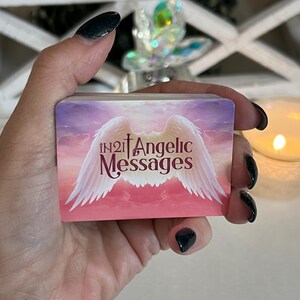 Puede incluir: Una baraja de cartas con un fondo rosa y morado que presenta un par de alas de &aacute;ngel blancas con una cruz y el texto "in2it Angelic Messages".