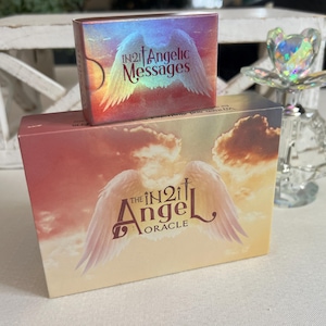 Puede incluir: Una caja de cartas de Oracle The IN2it Angel con un fondo rosa, naranja y amarillo con un par de alas de &aacute;ngel blancas. La caja tiene el texto "The IN2it Angel Oracle" impreso en ella. Una caja m&aacute;s peque&ntilde;a con el texto "IN2it Angelic Messages" est&aacute; encima de la caja m&aacute;s grande.