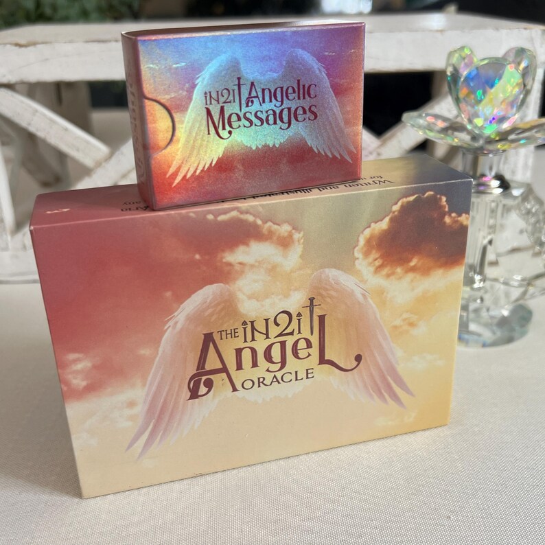 Puede incluir: Una caja de cartas de Oracle The IN2it Angel con un fondo rosa, naranja y amarillo con un par de alas de &aacute;ngel blancas. La caja tiene el texto "The IN2it Angel Oracle" impreso en ella. Una caja m&aacute;s peque&ntilde;a con el texto "IN2it Angelic Messages" est&aacute; encima de la caja m&aacute;s grande.