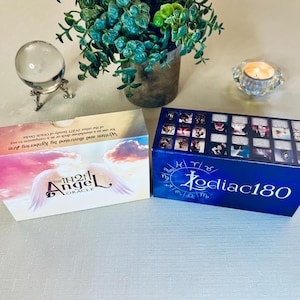 May include: Two boxes of oracle cards. The box on the left is white with a pink and purple sky and the words "The IN2IT Angel Oracle" in a gold font. The box on the right is blue with a white border and the words "Zodiac180" in a white font.