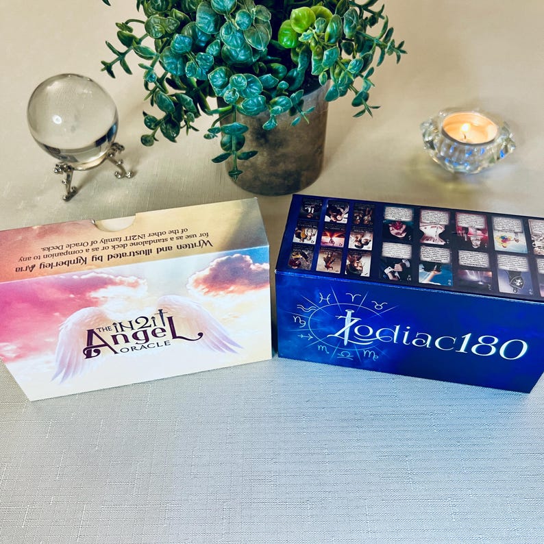 May include: Two boxes of oracle cards. The box on the left is white with a pink and purple sky and the words "The IN2IT Angel Oracle" in a gold font. The box on the right is blue with a white border and the words "Zodiac180" in a white font.