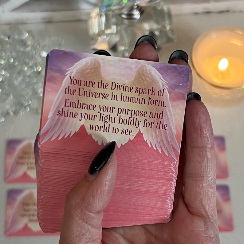 Puede incluir: Una tarjeta de afirmaci&oacute;n rosa y blanca con un mensaje sobre abrazar tu prop&oacute;sito y brillar tu luz. La tarjeta presenta un par de alas blancas y un cielo rosa y morado.