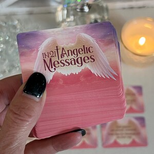 Puede incluir: Una baraja de cartas de mensajes angelicales con un fondo degradado rosa y morado. Las cartas presentan un dise&ntilde;o de alas de &aacute;ngel blancas y el texto "in2it Angelic Messages".