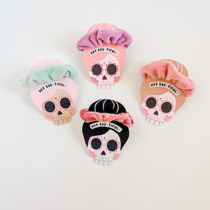 Op de afbeelding: Vier roze, bruine en zwarte suiker schedel Halloween decoraties met scrunchies erop. Elke schedel heeft een roze bloem ontwerp en de tekst "HEY BOO-TIFUL!" op de scrunchie.